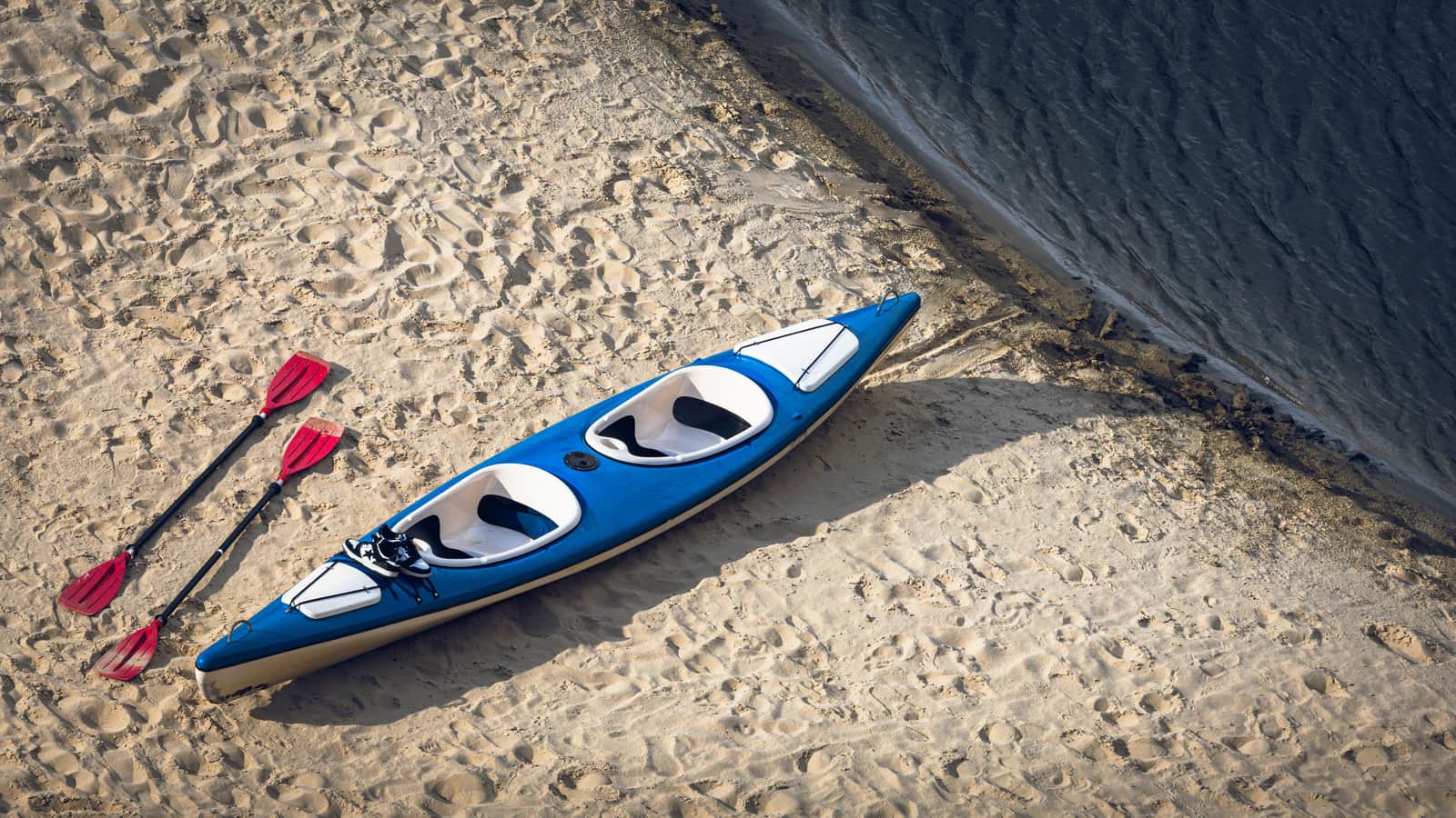A two-seat kayak and a pair of paddles resting on a sand beach at the water line