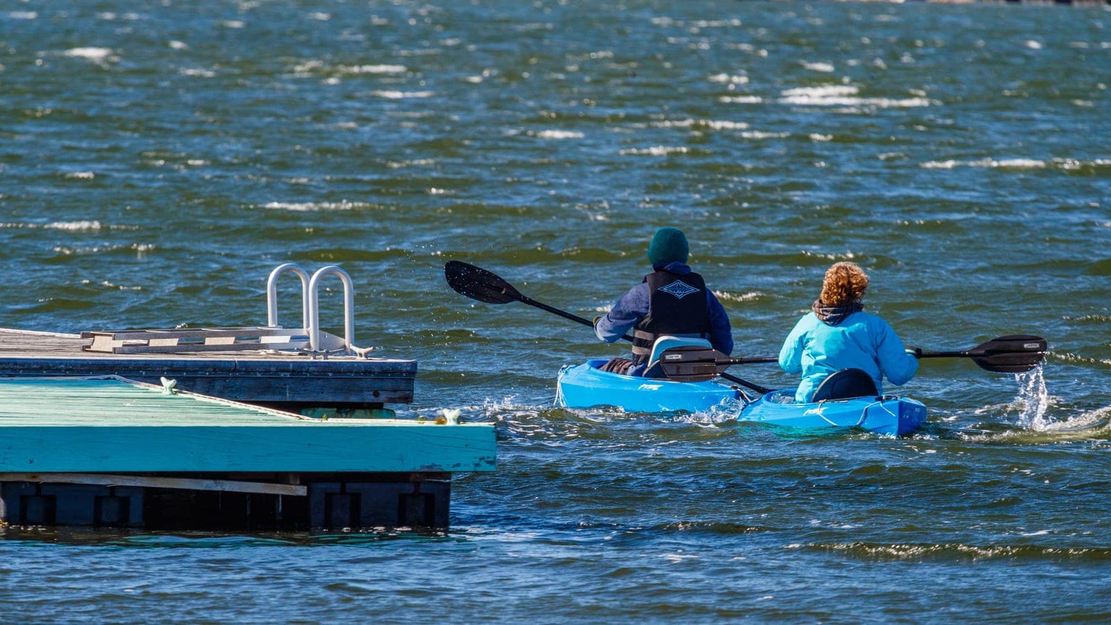 Two paddlers in separate kayaks setting out past a dock on open water