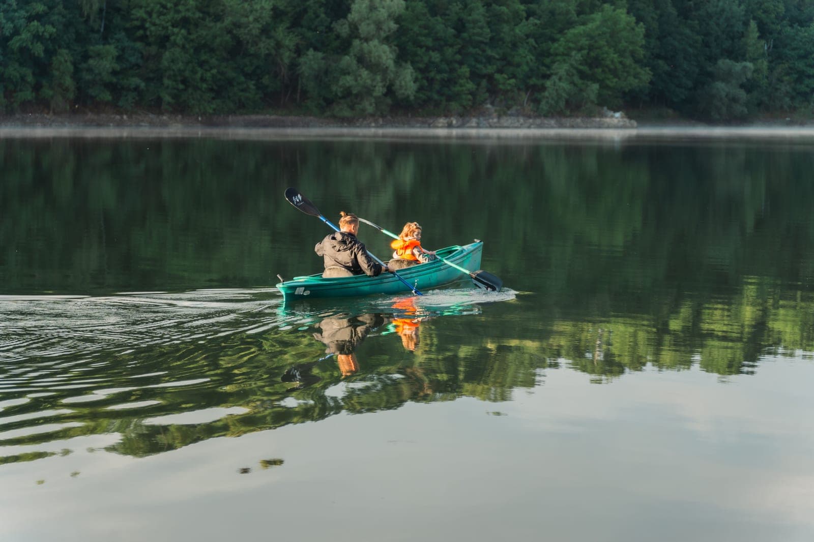 An adult and a child in a life jacket paddling a tandem kayak on a calm lake
